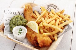 Fish and Chips in einer Schale, auf Zeitungspapier angerichtet. Dabei Erbsenpüree, Remoulade und Zitrone.