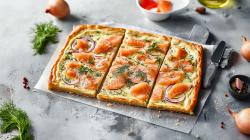 Ein Flammkuchen mit Räucherlachs, roten Zwiebeln und Dill, der auf einem Stück Backpapier auf einer grauen Arbeitsfläche liegt. Der Flammkuchen in drei Stücke geschnitten. Um den Flammkuchen herum liegen frischer Dill und Zwiebeln verteilt. Außerdem ist eine Schale mit Lachsstücken und eine Schale mit Knoblauchzehen zu sehen und im Hintergrund ein Gefäß mit Öl angeschnitten.