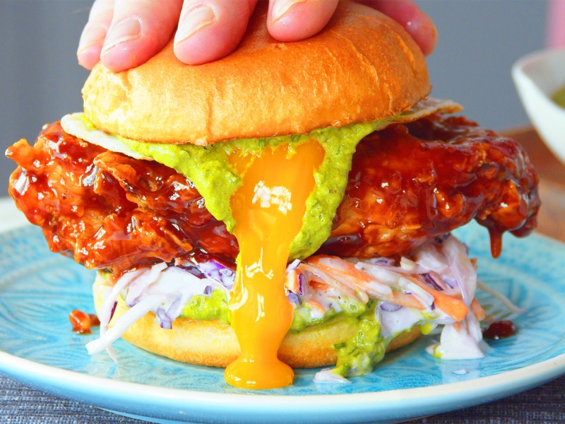 Ein Fried Chicken Burger auf einem blauen Teller wird leicht zerdrückt, sodass Soßen und Eigelb etwas aus ihm herauslaufen.