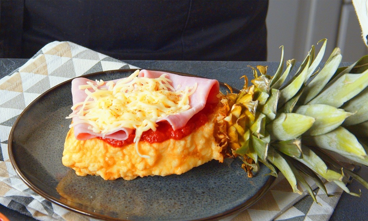 Ein Stück Ananas-Pizza, das aus einem mit Hefeteig ummantelten und frittierten Ananasstück besteht, welches mit Tomatensoße Schinken und Käse belegt ist. Der Strunk ist noch am Ananasstück dran. Die Ananas-Pizza liegt auf einem dunkelgrauen Teller, der auf einem gemusterten Küchentuch liegt.