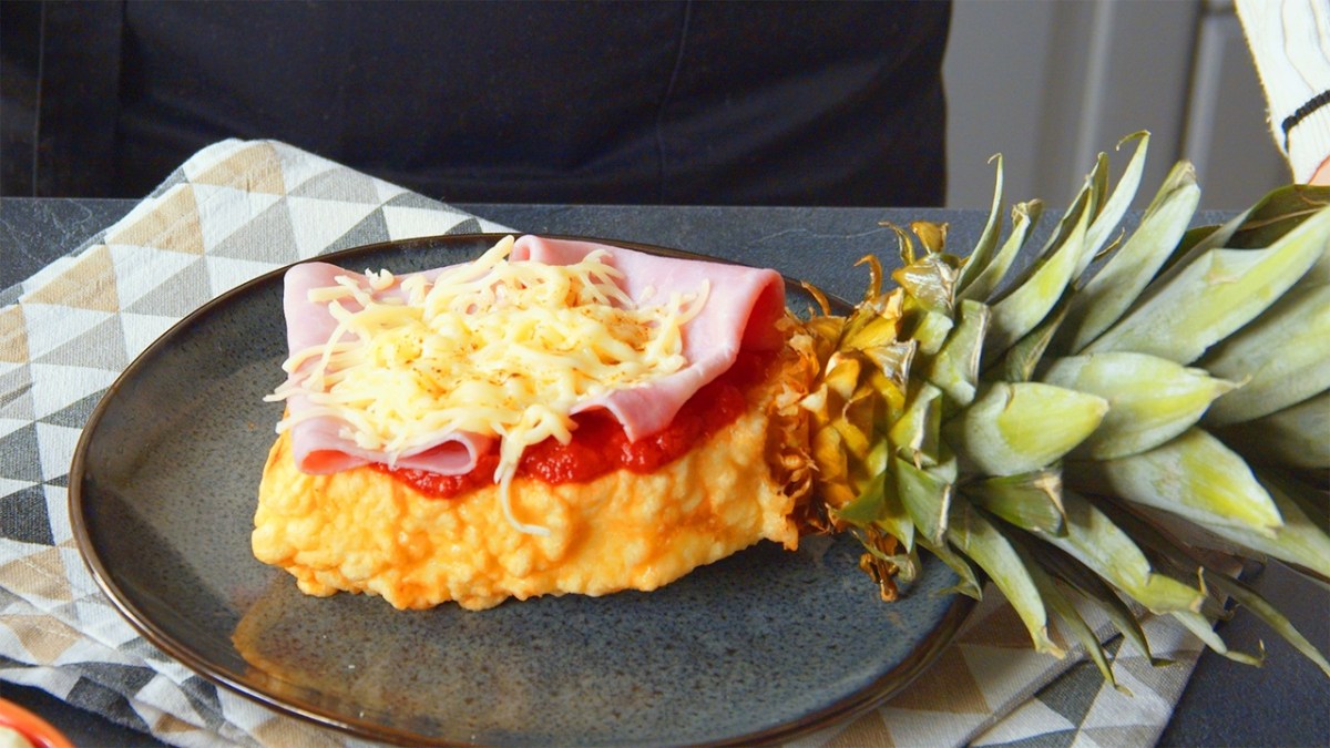 Ein Stück Ananas-Pizza, das aus einem mit Hefeteig ummantelten und frittierten Ananasstück besteht, welches mit Tomatensoße Schinken und Käse belegt ist. Der Strunk ist noch am Ananasstück dran. Die Ananas-Pizza liegt auf einem dunkelgrauen Teller, der auf einem gemusterten Küchentuch liegt.