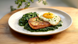 Ein großer Teller mit gebratenem Leberkäse mit Spiegelei und Spinat garniert mit etwas frischem Schnittlauch. Der Teller steht auf einem braunem Holztisch. Im Hintergrund sind Zweige einer Pflanze zu sehen.