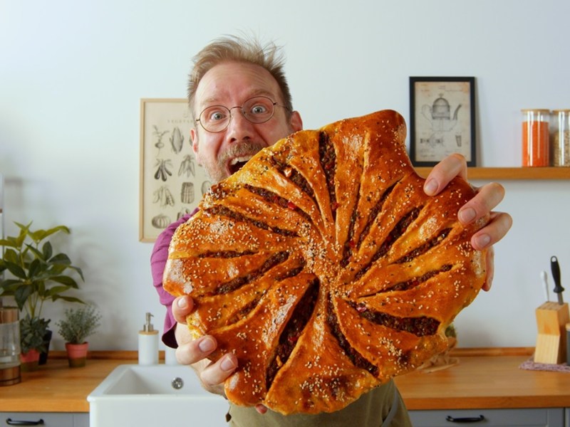 Koch Oli präsentiert ein großes selbst gemachtes und knusprig goldbraun gebackenes Hack-Fladenbrot. Im Hintergrund ist eine Küche zu sehen.