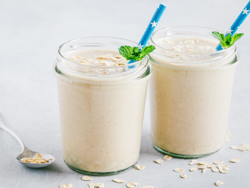 Zwei Gläser Käsekuchen-Shake mit blauen Strohhalmen und frischer Minze dekoriert. Die Käsekuchen-Shakes sind jeweils mit ein paar Haferflocken bestreut. Neben den Gläsern liegt ein Löffel mit Haferflocken und ringsum sind auch welche verstreut. Die Gläser stehen auf einer weißen Holzoberfläche.