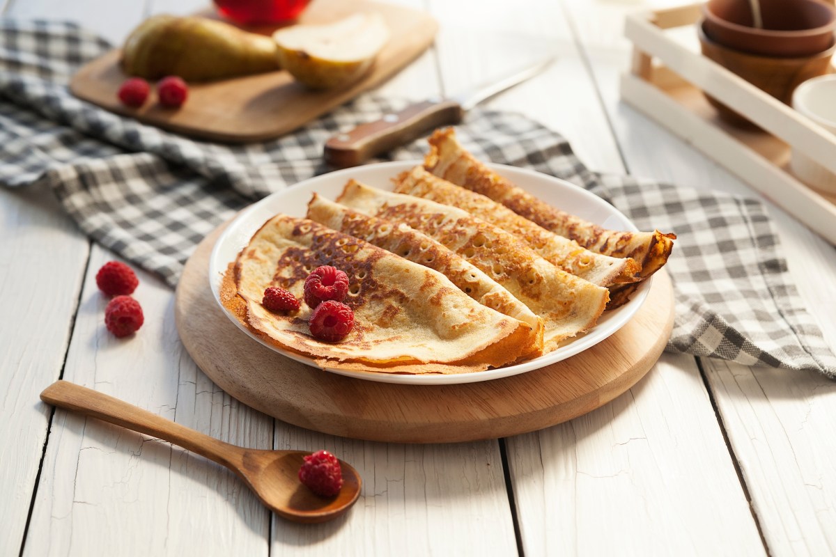 Einige Kefir-Pfannkuchen auf einem Teller, garniert mit Himbeeren, daneben einige Zutaten.