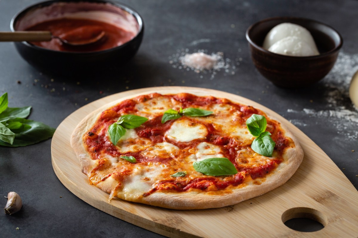 Eine Pizza Margherita mit Tomatensoße, Mozzarella und Basilikum auf einem großen Holzbrett, drumherum einige Zutaten