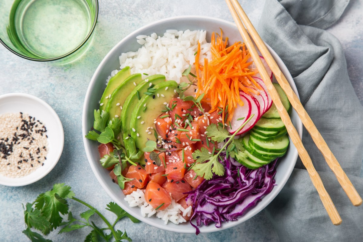 Poke Bowl mit frischem Fisch, Gemüse und Sesam in der Draufsicht. Daneben liegen einige der Zutaten.