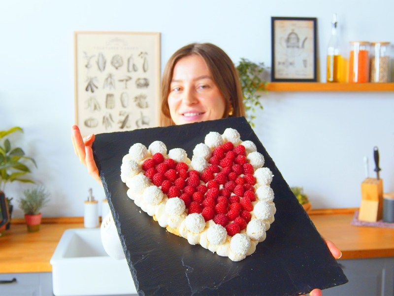 Unsere Köchin hält den Raffaello-Herzkuchen auf einer Schieferplatte in die Kamera. Der Raffaello-Herzkuchen besteht aus weichen Löffelbiskuits, ist am Rand von Raffaellos umgeben und mit Himbeeren dekoriert. Im Hintergrund sieht man eine Kochzeile mit, von links nach rechts, einem Wasserkocher, Spüle, einem Messerblock sowie Aufbewahrungsdosen.