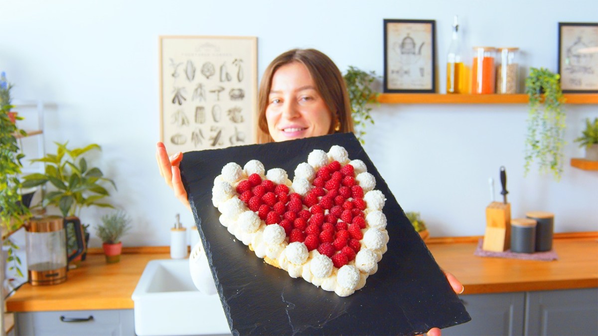 Unsere Köchin hält den Raffaello-Herzkuchen auf einer Schieferplatte in die Kamera. Der Raffaello-Herzkuchen besteht aus weichen Löffelbiskuits, ist am Rand von Raffaellos umgeben und mit Himbeeren dekoriert. Im Hintergrund sieht man eine Kochzeile mit, von links nach rechts, einem Wasserkocher, Spüle, einem Messerblock sowie Aufbewahrungsdosen.