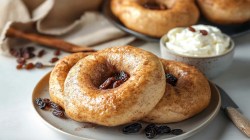 Ein Teller mit 3 Rosinen-Zimt-Bagel, daneben ein Messer und eine Schale Frischkäse, dahinter weitere Bagel.