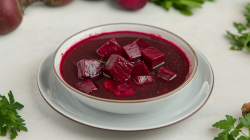 Eine Schüssel mit polnischer Roter-Bete-Suppe steht auf einem Unterteller auf einem weißen Tisch. In der Rote-Bete-Suppe befinden sich Rote-Bete-Würfel. Am Rand des Bildes sieht man einige frische Kräuter liegen, sowie hinten links eine ungeschälte rote Bete.