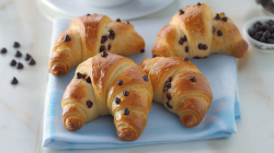 Vier Schokohörnchen, die auf einem hellblauen Tuch auf einer hellgrauen Arbeitsfläche liegen. Rechts im Hintergrund kann man eine kleine Schale mit Schokotropfen sehen. Links neben den Schokohörnchen liegen ein paar einzelne Schokotropfen. Hinter den Schokohörnchen steht noch ein leerer Teller.