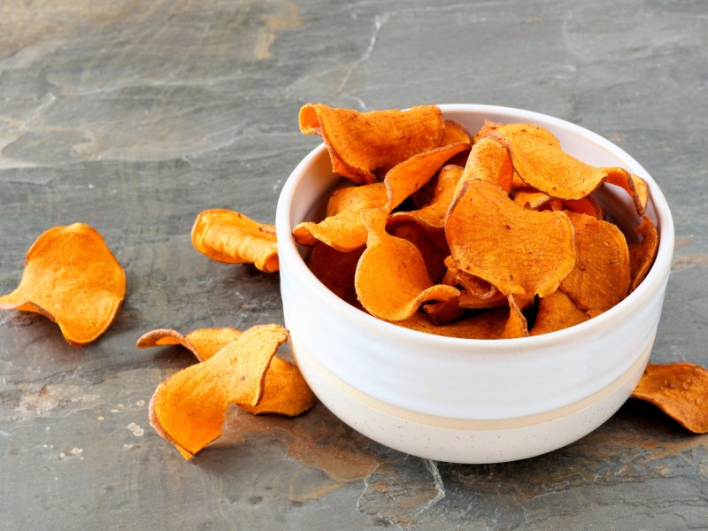 Eine weiße kleine Schüssel mit Süßkartoffel-Chips auf einer grauen Steinoberfläche. Ringsum die Schüssel liegen einzelne Süßkartoffel-Chips verstreut.