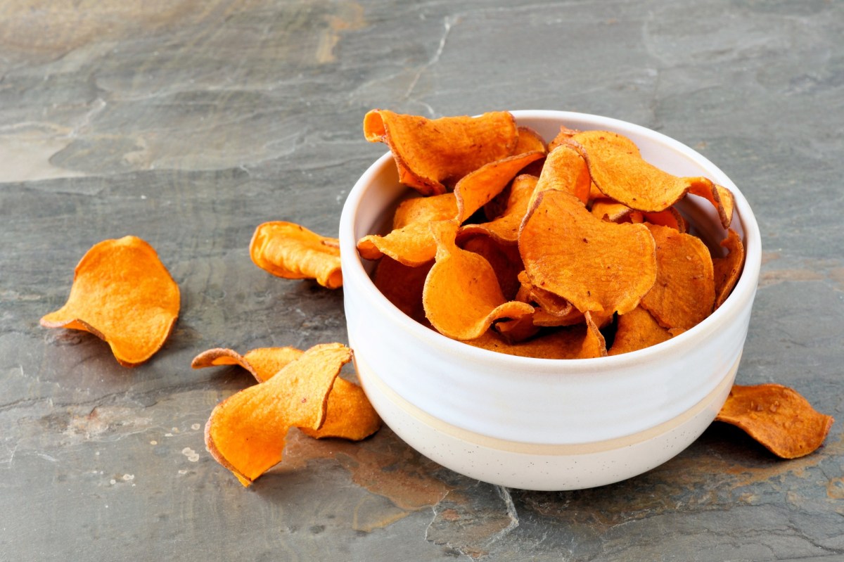 Eine weiße kleine Schüssel mit Süßkartoffel-Chips auf einer grauen Steinoberfläche. Ringsum die Schüssel liegen einzelne Süßkartoffel-Chips verstreut.