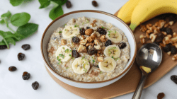 Eine Schüssel mit Zoats steht auf einem Holzbrett auf einem weißen Tisch. Recht daneben liegt ein Löffel, Nüsse, Rosinen und zwei Bananen. Links neben der Schüssel liegen weitere Rosinen und ein wenig frische Kräuter zu Dekoration.