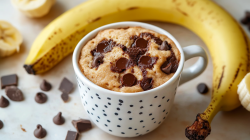 Ein Bananen-Tassenkuchen steht auf einem hellen Untergrund. Um die Tasse verteilt liegen Schokoladenstücke und eine Banane.