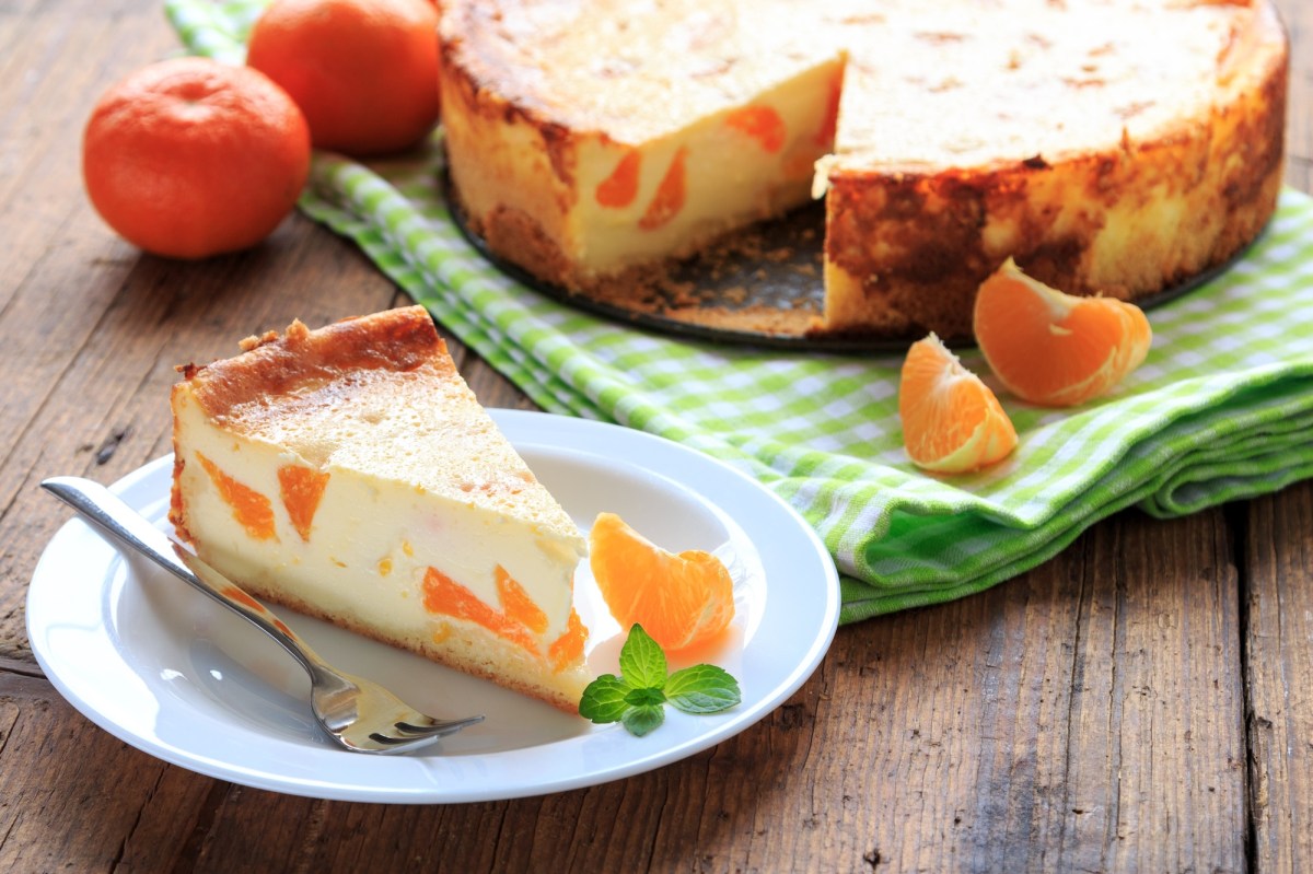 Ein Stück Mandarinen-Quark-Kuchen auf einem Teller, daneben der Kuchen und ein paar Mandarinen.