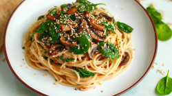 Sesam-Pilz-Spaghetti auf einem Teller. Sie sind mit Champignons, Spinat und Sesamkörnern bestreut. Rechts neben dem Teller liegen einige weitere Spinatblätter.