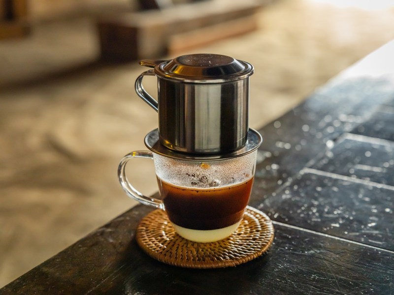 Ein Glas vietnamesischer Kaffee mit Filter steht auf einem schwarzen Holztisch. Man kann deutlich die Kondensmilch im Kaffee erkennen.